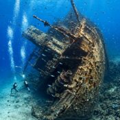 Scuba divers exploring a large tilted shipwreck covered in coral and surrounded by fish in the Red Sea
